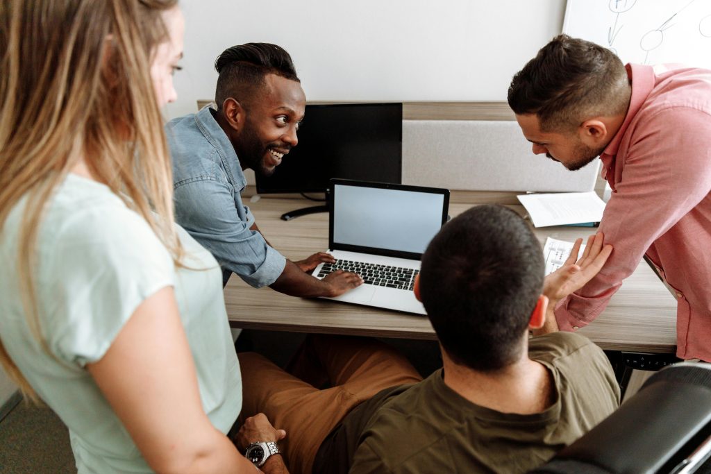 Quatre collègues discutent autour d’un ordinateur portable sur un bureau, en réunion d’équipe.